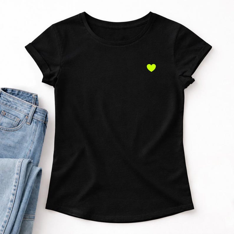 Może przedstawiać: Czarny t-shirt z okrągłym dekoltem i małym neonowo-zielonym sercem na lewej piersi. T-shirt jest połączony z niebieskimi dżinsami. Koszulka ma kr&oacute;tkie rękawy i klasyczny, swobodny styl.