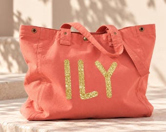 May include: A coral-colored canvas tote bag featuring the letters ILY in a gold glitter finish on the front. The bag includes two top handles, reinforced stitching, and metallic grommets at the top corners for a structured and durable design.