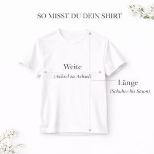 Może przedstawiać: Biała koszulka z tekstem i strzałkami wskazującymi, jak zmierzyć koszulkę. Tekst brzmi "SO MISST DU DEIN SHIRT", "Weite (Achsel zu Achsel)" i "L&auml;nge (Schulter bis Saum)". Koszulka jest wyświetlana na jasnym tle z kwiatowymi akcentami.