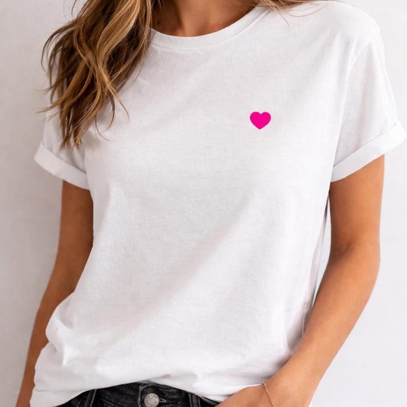 Könnte beinhalten: Ein weißes T-Shirt mit kurzen Ärmeln und gerolltem Saum. Auf der linken Brust ist ein kleines, leuchtend pinkfarbenes Herz aufgestickt. Das Shirt besteht aus einem weichen, leichten Material.