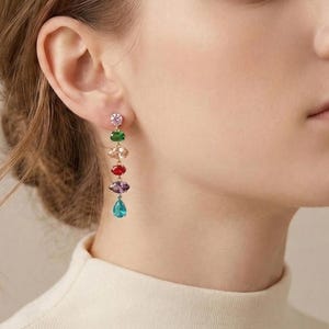 May include: A colorful dangle earring with a series of gemstones in pink, green, gold, red, purple, and teal. The earring is worn on the ear of a person with light skin and brown hair. The earring is a statement piece.