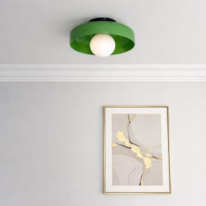 May include: A modern ceiling light fixture with a green shade and a white globe bulb. The light is mounted on a black base. A framed abstract art print with gold accents hangs below.