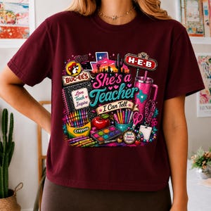 May include: A maroon t-shirt featuring a colorful graphic print with the text 'She's a Teacher I Can Tell', 'Buc-ee's', 'H-E-B', 'Love Teach Inspire', and 'OMG'. The design includes illustrations of crayons, a notebook, an apple, a tumbler, and scissors.