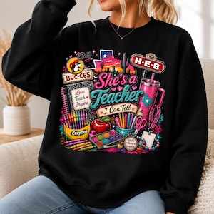 May include: A black crewneck sweatshirt featuring a colorful graphic print with the text "She's a Teacher I Can Tell," "Buc-ee's," "H-E-B," "Love Teach Inspire," and "Crayons." The design includes a Texas outline, a tumbler, and school supplies.