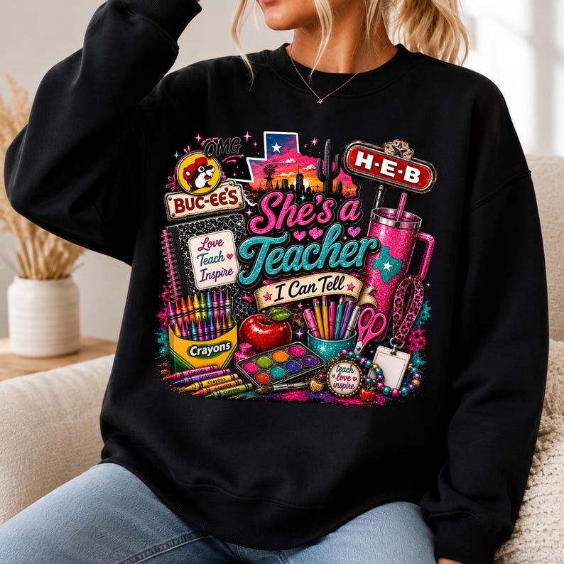 May include: A black crewneck sweatshirt featuring a colorful graphic print with the text "She's a Teacher I Can Tell," "Buc-ee's," "H-E-B," "Love Teach Inspire," and "Crayons." The design includes a Texas outline, a tumbler, and school supplies.