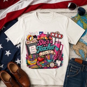 May include: A cream-colored t-shirt featuring a colorful graphic print with the text 'She's a Teacher I Can Tell', 'Buc-ee's', 'H-E-B', and 'Love Teach Inspire'. The design includes illustrations of crayons, a red apple, a tumbler, and school supplies.