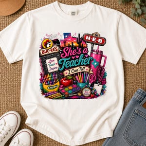 May include: A white t-shirt featuring a colorful graphic print with the text "She's a Teacher I Can Tell". The design includes a Buc-ee's logo, an H-E-B sign, a box of crayons, a red apple, a pink tumbler, scissors, paint, and school supplies.