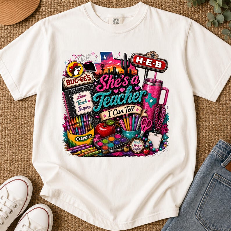 May include: A white t-shirt featuring a colorful graphic print with the text "She's a Teacher I Can Tell". The design includes a Buc-ee's logo, an H-E-B sign, a box of crayons, a red apple, a pink tumbler, scissors, paint, and school supplies.