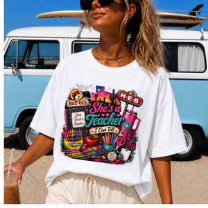 May include: A white t-shirt featuring a colorful graphic print with the text "She's a Teacher I Can Tell". The design includes a Buc-ee's logo, an H-E-B sign, a pink tumbler, a red apple, crayons, a watercolor paint set, and a notebook.