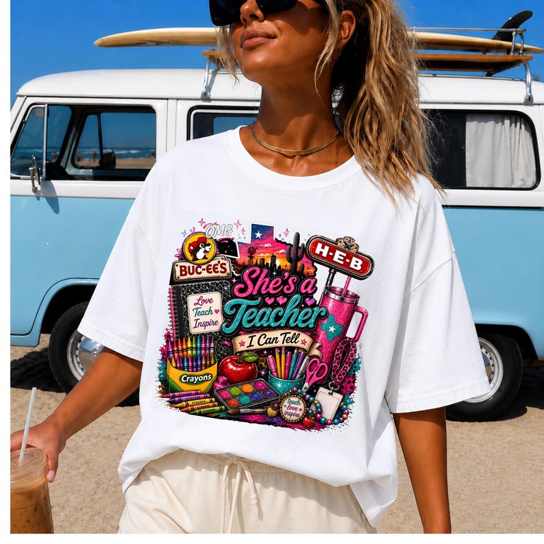 May include: A white t-shirt featuring a colorful graphic print with the text "She's a Teacher I Can Tell". The design includes a Buc-ee's logo, an H-E-B sign, a pink tumbler, a red apple, crayons, a watercolor paint set, and a notebook.