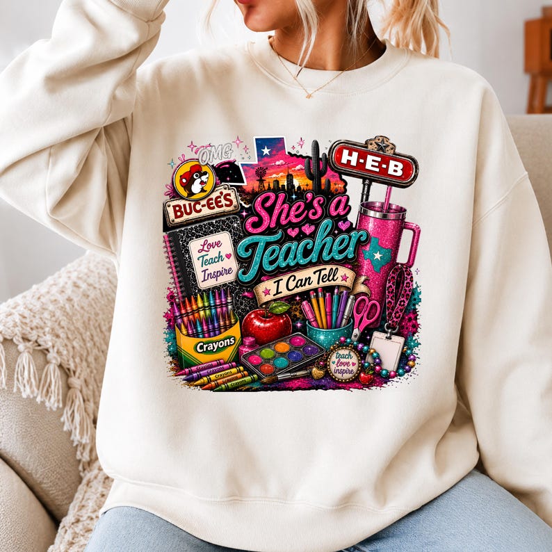 May include: A cream-colored crewneck sweatshirt featuring a colorful graphic print with the text "She's a Teacher I Can Tell," "Buc-ee's," "H-E-B," and "Love Teach Inspire." The design includes school supplies like crayons, a notebook, an apple, and a tumbler.