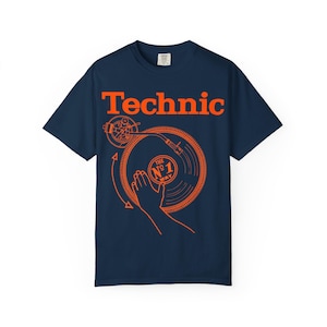 Puede incluir: Camiseta azul marino con la palabra "Technic" en naranja sobre una ilustración naranja de un tocadiscos. El gráfico del tocadiscos incluye una mano, un disco y el texto "The No. 1 Way."