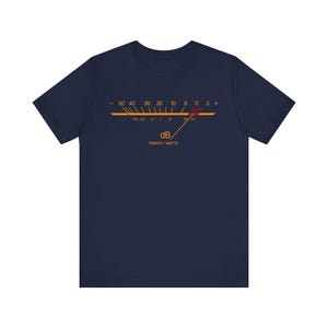 Puede incluir: Una camiseta azul marino con un estampado gráfico de un medidor de decibelios analógico. El diseño incluye una escala horizontal con números del -50 al +3, una aguja que apunta al cero y el texto dB y POWER / WATTS en color naranja.
