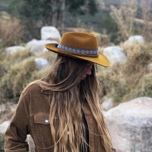 Könnte beinhalten: Senffarbener Fedora-Hut mit einem dekorativen Band mit einem geometrischen Muster in Weiß und Schwarz. Der Hut hat eine breite Krempe und eine geflochtene Bordüre. Die Person trägt eine braune Cordjacke.