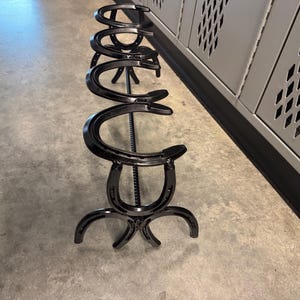 May include: A black metal horseshoe boot rack. The rack is made of multiple horseshoes welded together. The rack is approximately 36 inches tall and 18 inches wide.