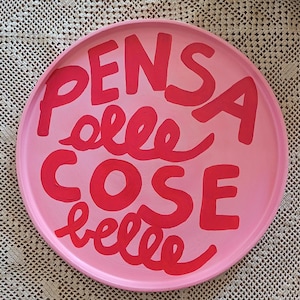 Könnte beinhalten: Ein runder, rosa dekorativer Teller mit den Worten "PENSA cose belle" in roter Schrift. Der Teller hat eine glatte Oberfläche und einen leicht erhöhten Rand. Der Text ist in einem verspielten, handschriftlichen Stil gehalten.
