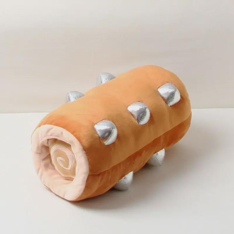 May include: A plush, cylindrical toy in the shape of a Swiss roll cake. The toy is primarily orange with silver accents and a cream-colored swirl visible at one end. The plush toy is designed to resemble a dessert.