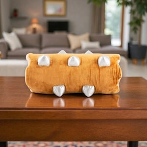 May include: A plush, mustard-yellow pillow shaped like a log, featuring six silver, cone-shaped accents. The pillow is rectangular and sits on a dark wooden table, with a blurred living room in the background.