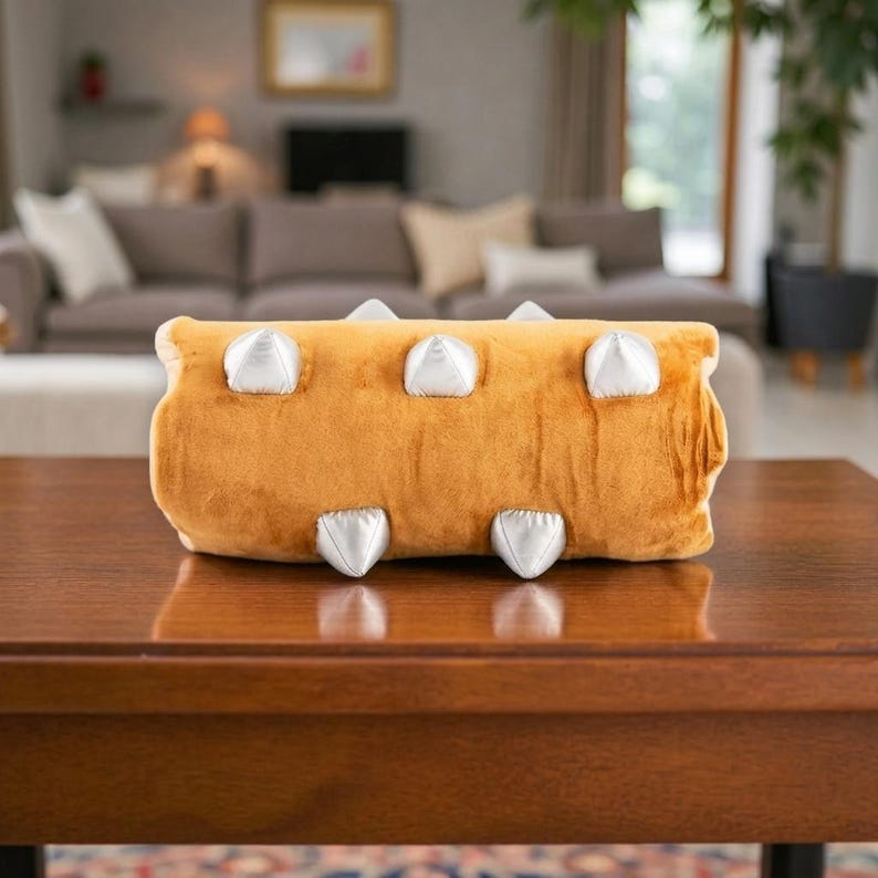 May include: A plush, mustard-yellow pillow shaped like a log, featuring six silver, cone-shaped accents. The pillow is rectangular and sits on a dark wooden table, with a blurred living room in the background.