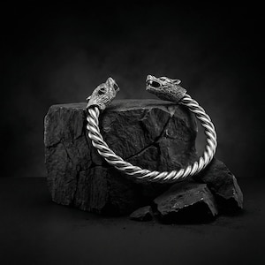 Puede incluir: Un brazalete plateado con terminales de cabeza de lobo. El brazalete tiene un diseño de cuerda trenzada y se exhibe sobre una roca oscura y texturizada. Las cabezas de lobo están detalladas con bocas abiertas y patrones de pelaje intrincados.