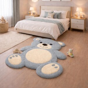 Pode incluir: Um tapete grande, azul, em forma de urso de pelúcia, com um centro e almofadas das patas creme, está sobre um chão de madeira num quarto. O tapete é rodeado por uma cama com roupa de cama branca, uma manta azul clara e almofadas a condizer.