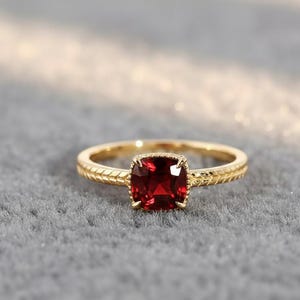 Könnte beinhalten: Ein goldener Ring mit einem quadratischen, tiefroten Edelstein. Das Band hat ein strukturiertes Design. Der Ring ist vor einem weichen, grauen Hintergrund platziert, der die Details und die Farbe des Schmucks hervorhebt.