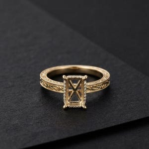 Könnte beinhalten: Ein goldener Ring mit einer rechteckigen Fassung, die von einem feinen Diamantbesatz umrandet ist. Die Ringschiene ist an den Seiten mit detaillierten, eingravierten Rankenmustern verziert, die der Metalloberfläche eine Struktur verleihen.