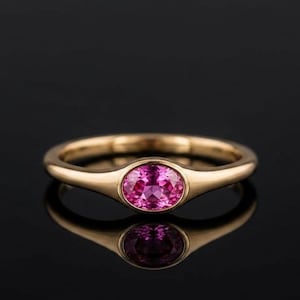 Könnte beinhalten: Ein goldener Ring mit einem einzelnen, oval geschliffenen, leuchtend rosa Edelstein in einer glatten Zargenfassung. Die Ringschiene ist poliert und abgerundet und zeigt eine reflektierende Oberfläche, die die magentafarbenen Facetten betont.