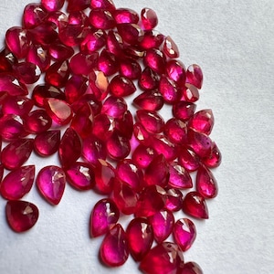 Könnte beinhalten: Eine Ansammlung facettierter, tropfenförmiger Rubine in verschiedenen Größen. Die Edelsteine sind tiefrot und haben einen hohen Reinheitsgrad, der das Licht reflektiert. Die Rubine sind auf einer weißen Oberfläche verstreut.