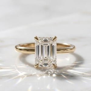 Könnte beinhalten: Ein goldener Verlobungsring mit einem großen, rechteckigen Diamanten im Smaragdschliff. Der klare, facettierte Diamant ist in einem schlichten Goldband gefasst. Der Ring wird auf einer weißen Oberfläche mit Lichtreflexen präsentiert.
