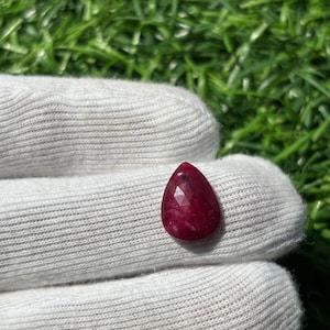 Könnte beinhalten: Ein facettierter, tropfenförmiger Rubin-Edelstein. Der Rubin hat eine tiefe, satte rote Farbe und wird von einer behandschuhten Hand gehalten. Der Hintergrund ist verschwommen und zeigt grünes Gras.