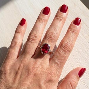 Könnte beinhalten: Ein Silberring mit einem großen, oval geschliffenen, tiefroten Edelstein in einer glatten Metallfassung. Der Ring wird an einem Finger getragen und zeigt die facettierte Oberfläche des Steins, die das Licht reflektiert und kräftig leuchtet.