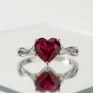 Könnte beinhalten: Ein silberner Ring mit einem herzförmigen, tiefroten Edelstein. Der Ring hat ein verdrehtes Banddesign mit kleinen Akzentsteinen auf beiden Seiten des herzförmigen Steins. Der Ring wird auf einer reflektierenden Oberfläche präsentiert.
