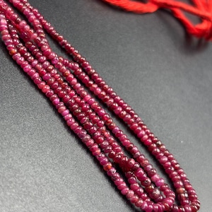 Puede incluir: Primer plano de múltiples hilos de pequeñas gemas redondas de color rojo intenso, posiblemente rubíes, sobre un fondo oscuro. En la parte superior se ve un hilo rojo brillante, lo que sugiere un collar o una joya. Las gemas tienen un aspecto pulido.