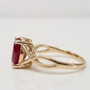 Könnte beinhalten: Ein Goldring mit einem facettierten, oval geschliffenen, tiefroten Edelstein in einer Vier-Krappen-Fassung. Die Ringschiene besteht aus poliertem Gelbgold und zeigt ein offenes, gespaltenes Design, das beidseitig in die Fassung übergeht.
