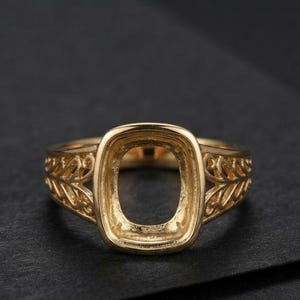 Könnte beinhalten: Ein goldfarbener Ringrohling mit einer zentralen, rechteckigen Fassung mit abgerundeten Ecken zur Aufnahme eines Edelsteins. Die Ringschiene zeigt ein filigranes, durchbrochenes Blatt- oder Rankenmuster, das sich seitlich erstreckt.