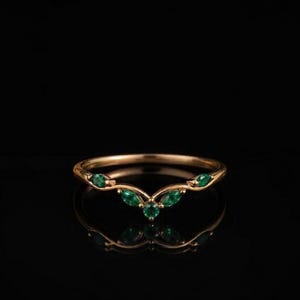 Könnte beinhalten: Ein goldener V-förmiger Ring mit fünf kleinen, runden und marquise-geschliffenen grünen Edelsteinen entlang der Ringschiene. Der Ring hat eine polierte metallische Oberfläche und ein geschwungenes Design, das in einem zentralen Punkt zuläuft.