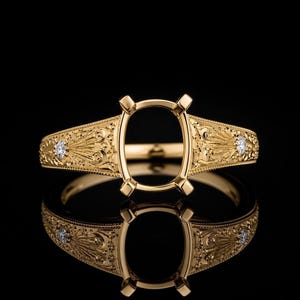 Könnte beinhalten: Ein goldener Ringrohling mit einer ovalen Fassung und vier Krappen. Die Ringschiene ist mit detaillierten Gravuren verziert und auf beiden Seiten der zentralen Fassung mit jeweils einem kleinen, runden, klaren Stein besetzt.