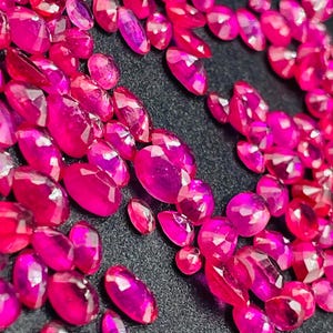 Può includere: Una collezione di gemme di rubino sfaccettate, di forma ovale e rotonda. Le gemme sono di un vibrante colore rosa-rosso e sono sparse su una superficie scura. Le gemme variano in dimensioni e forma, riflettendo la luce.