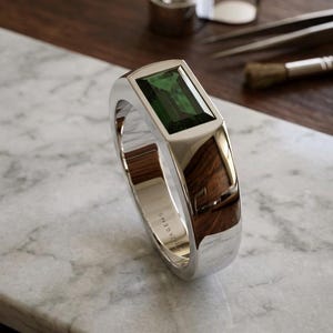 Könnte beinhalten: Ein silberner Ring mit einem rechteckigen, smaragdgrünen Edelstein. Der Ring hat eine polierte Oberfläche und eine Holzeinlage. Die Worte "SWEDEGEMS" sind auf der Seite eingraviert. Er wird auf einer Marmoroberfläche präsentiert.