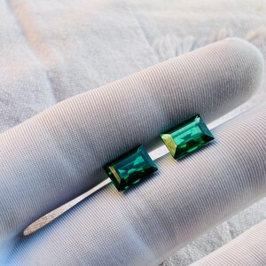 Könnte beinhalten: Zwei smaragdgrüne Edelsteine im Emerald-Schliff. Die rechteckigen Edelsteine haben eine facettierte Oberfläche und zeigen einen satten, tiefgrünen Farbton. Sie sind vor einem weißen Hintergrund platziert, was ihre Farbe und Klarheit hervorhebt. Die Edelsteine sind wahrscheinlich für Schmuck.
