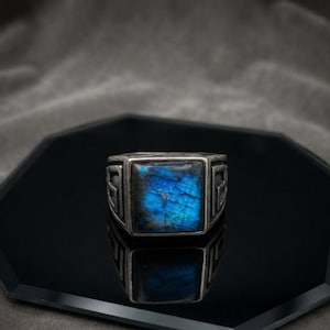 Puede incluir: Un anillo de tono plateado con una piedra de labradorita de corte cuadrado que muestra un destello azul iridiscente. La banda metálica presenta un patrón geométrico y angular en los laterales, enmarcando la piedra central en un engaste sólido.