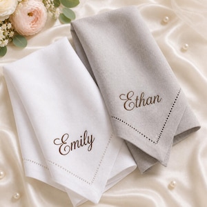 May include: Two folded fabric napkins, one white and one light gray, each featuring a decorative hemstitch border and personalized embroidery in a dark brown script font. The white napkin displays the name Emily, and the gray napkin displays Ethan.