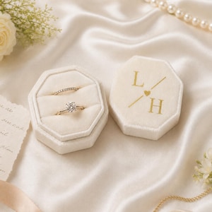 Puede incluir: Un joyero octogonal de terciopelo crema con dos anillos, uno con una piedra solitaria redonda y otro con una banda pavé. La tapa presenta letras doradas L / H con un pequeño corazón. El joyero está colocado sobre una superficie de satén crema.