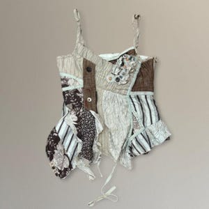 May include: A sleeveless camisole top constructed from a patchwork of fabrics in shades of brown, beige, and white. The garment features thin shoulder straps, a mix of striped and floral patterns, textured panels, and a cluster of decorative buttons.