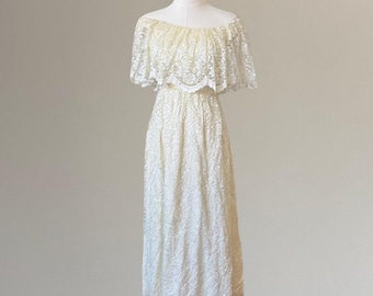 Puede incluir: Un vestido largo de color crema con hombros descubiertos y un patrón de encaje floral en toda la prenda. Incluye un volante ancho en el escote que cubre los hombros y una falda de corte recto que llega hasta la altura de los tobillos.