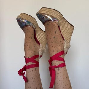 May include: A pair of wedge espadrilles featuring a clear, iridescent toe box and a woven jute platform sole. The shoes are secured with bright red ribbon ankle ties that wrap around the lower leg, worn over sheer tights with a small star pattern.