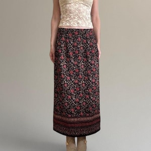 May include: A floor-length black skirt featuring a dense pattern of small red flowers and thin green stems. The hem is finished with a wide, horizontal band containing intricate, repeating geometric and floral motifs in red and black.