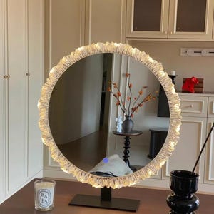 Op de afbeelding: Een ronde make-upspiegel met een getextureerde, verlichte rand. De spiegel staat op een zwarte standaard en weerspiegelt een kamer met een tafel, vaas en kaars. De rand van de spiegel is verlicht met kleine lampjes.