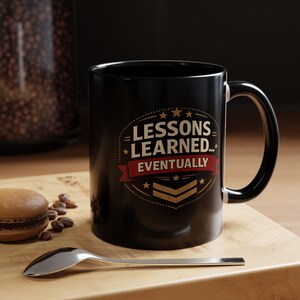 May include: Black ceramic mug with the text "LESSONS LEARNED... EVENTUALLY" in a white and red design. The mug has a black handle and is sitting on a wooden surface. A spoon and a macaron are also visible.